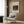 May include: Framed art print of Henri Rousseau's "The Rabbit's Meal" showing a gray and white rabbit eating orange carrots beside green cabbage leaves against a brick wall background. The cream-colored poster with black text hangs on wood-paneled wall above a beige upholstered armchair with geometric patterned pillow. Modern brass floor lamp and dark round side table with open book complete the neutral-toned reading nook scene.
