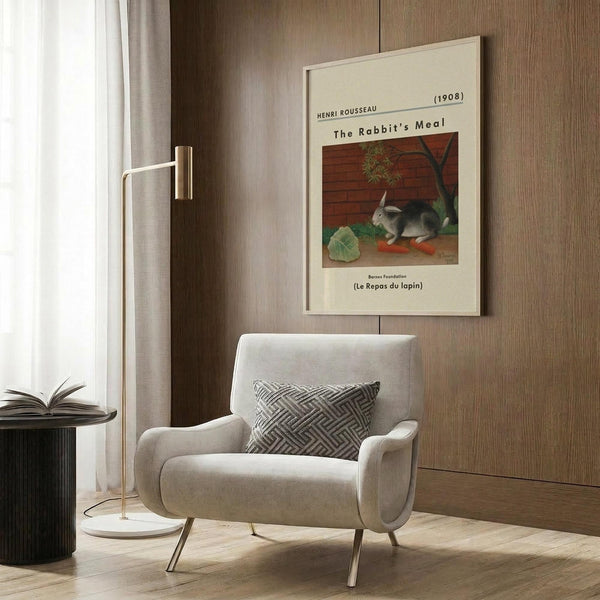 May include: Framed art print of Henri Rousseau's "The Rabbit's Meal" showing a gray and white rabbit eating orange carrots beside green cabbage leaves against a brick wall background. The cream-colored poster with black text hangs on wood-paneled wall above a beige upholstered armchair with geometric patterned pillow. Modern brass floor lamp and dark round side table with open book complete the neutral-toned reading nook scene.