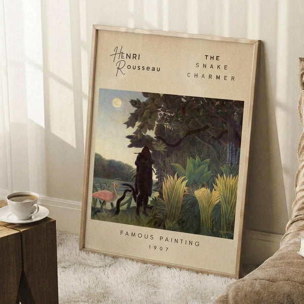 May include: Framed art print leaning against white wall on white textured rug, showing tropical jungle scene with silhouetted figure playing flute among lush green foliage, flamingo, and full moon in pale sky. Cream-colored mat with black text. White coffee cup on wooden block nearby, beige throw blanket visible at edge.