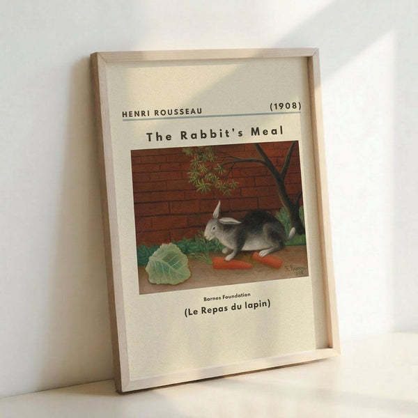 May include: Framed reproduction of Henri Rousseau's 1908 painting showing a gray and white rabbit sitting beside orange carrots and green cabbage leaves against a brick wall background. The museum-style print features cream matting with black text identifying the artist, title, and date. The light wood frame leans against a white wall, casting soft shadows.