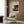 May include: Henri Rousseau's The Snake Charmer vintage art poster displayed in a modern living room setting with warm wood paneling. The framed print features a jungle scene with dark silhouetted figure, lush tropical plants, and moonlit sky in muted greens and blues. Shown above a cream upholstered armchair with geometric patterned throw pillow, beside a brass floor lamp and dark round side table with open book.