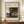 May include: Framed art print displayed on wooden credenza in neutral room setting. Print features jungle scene with silhouetted figure playing flute near water, surrounded by lush tropical foliage, pink flamingo, and full moon in dusky sky. Cream matting with brown frame. White ceramic vase and stacked books nearby.