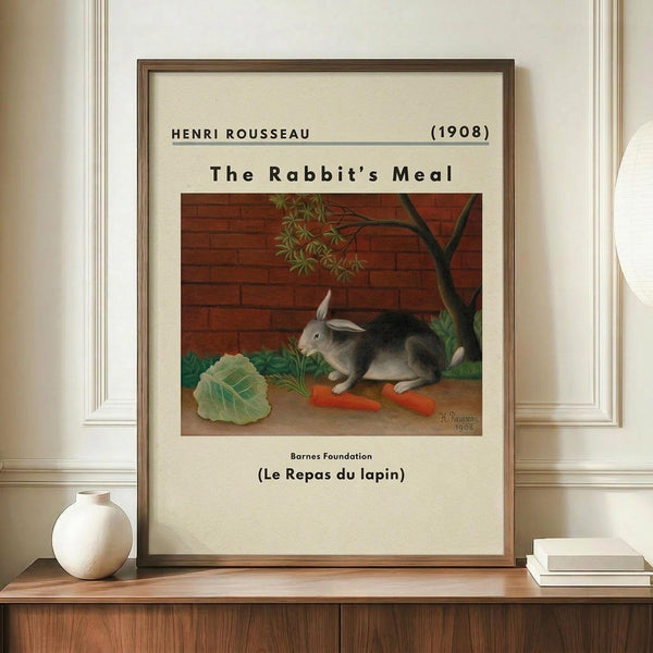 May include: Framed art print displaying Henri Rousseau's 1908 painting of a gray rabbit sitting beside orange carrots and green cabbage against a red brick wall with tree branches. The cream-colored poster shows museum-style text above and below the artwork. Displayed on wooden sideboard with white ceramic vase and stacked books in bright room with white wainscoting.