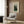 May include: Framed Henri Rousseau art print displayed on wood-paneled wall above cream upholstered armchair with geometric patterned pillow. The vintage-style poster features two figures in dark clothing against a lush green landscape with trees and foliage. Modern brass floor lamp with white base stands nearby next to dark round side table with open book. Soft natural lighting from window with sheer curtains.