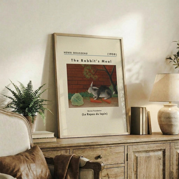 May include: Framed art print displayed on rustic wooden sideboard in neutral-toned living room. Print shows rabbit with vegetables against brick wall background, with cream matting and light wood frame. Scene includes fern plant, stacked books, ceramic vase, and beige upholstered furniture with brown throw pillows.