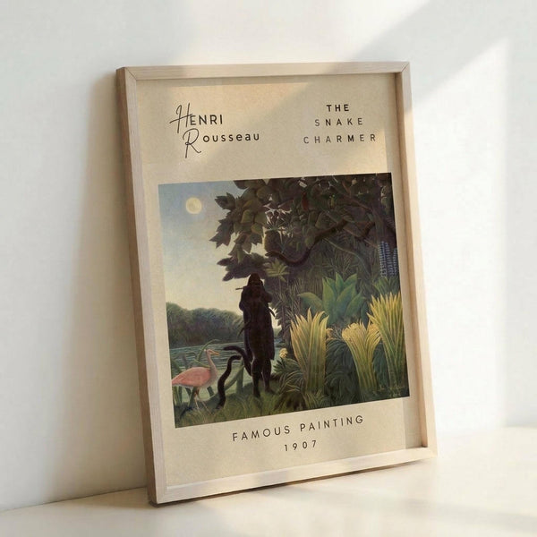 May include: Framed art print leaning against white wall showing jungle scene with silhouetted figure playing flute among lush tropical plants, pink flamingo, and full moon. Cream-colored poster with text identifying Henri Rousseau's "The Snake Charmer" from 1907. Natural wood frame, neutral background lighting creates soft shadows.