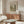 May include: Framed vintage-style landscape print hanging above wooden sideboard in minimalist living room. Print shows pastoral scene with trees and water in muted green and beige tones. Room features cream walls, white upholstered chair, glass oval coffee table, and copper floor lamp. Natural lighting creates warm, serene atmosphere.