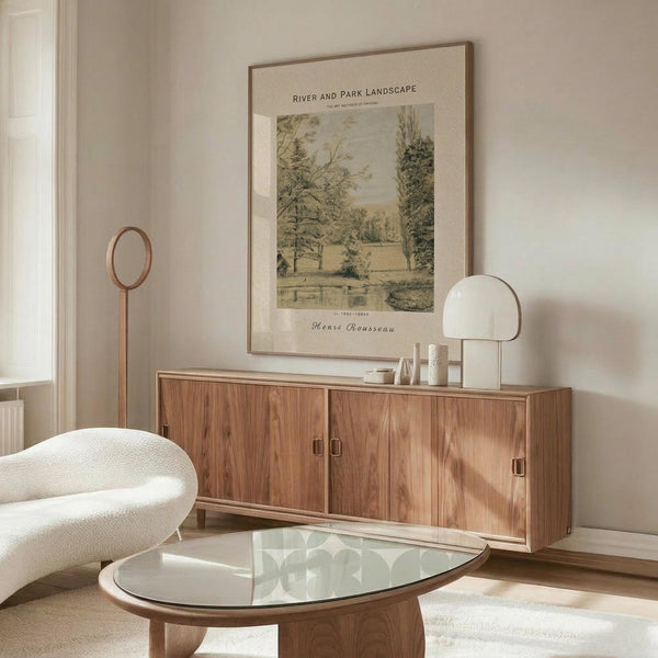May include: Framed vintage-style landscape print hanging above wooden sideboard in minimalist living room. Print shows pastoral scene with trees and water in muted green and beige tones. Room features cream walls, white upholstered chair, glass oval coffee table, and copper floor lamp. Natural lighting creates warm, serene atmosphere.