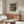 May include: Framed vintage art print displayed on wooden credenza in modern living room setting. Print features muted green and beige landscape scene. Room styled with cream upholstered chair, glass-top oval coffee table, sculptural floor lamp, and table lamp. Warm wood tones throughout with neutral color palette and natural lighting from window.