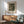 May include: Framed vintage art print displayed on white wall above wooden sideboard in modern living room. The artwork shows a muted green and brown landscape scene from 1865 with trees and buildings. Mid-century modern sage green armchair with wooden frame sits beside the credenza, along with white ceramic table lamp and round wooden coffee table. Warm natural sunlight creates geometric shadows across the scene.
