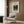 May include: Henri Rousseau jungle artwork print in cream mat and frame, mounted on wood-paneled wall above beige upholstered armchair with geometric patterned pillow. Golden brass floor lamp with white marble base stands nearby next to dark round side table with open book. Soft natural lighting from window with sheer curtains illuminates the modern neutral-toned interior setting.