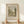 May include: Two hands holding a framed vintage landscape print showing trees, water, and parkland in muted green and beige tones. The artwork features "River and Park Landscape" text and "Henri Rousseau" attribution below the image. Light wood frame sits on a white wooden surface against a cream wall, with soft natural lighting creating gentle shadows.