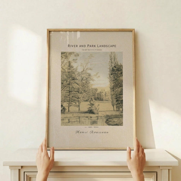 May include: Two hands holding a framed vintage landscape print showing trees, water, and parkland in muted green and beige tones. The artwork features "River and Park Landscape" text and "Henri Rousseau" attribution below the image. Light wood frame sits on a white wooden surface against a cream wall, with soft natural lighting creating gentle shadows.