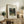 May include: Framed botanical art print featuring jungle scene with monkeys and colorful parrot among lush green foliage, displayed on rustic wooden sideboard alongside fern plant, ceramic vase, and books in neutral-toned living room setting with cream upholstered furniture.