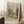 May include: A framed vintage-style landscape print leaning against a white wall corner, featuring trees, a river, and pastoral scenery in sepia tones. The print shows Art Institute of Chicago attribution and Henri Rousseau signature at bottom. A wooden side table holds a white coffee cup, with a cream knit throw blanket nearby on white carpet. Natural sunlight creates soft shadows on the wall.