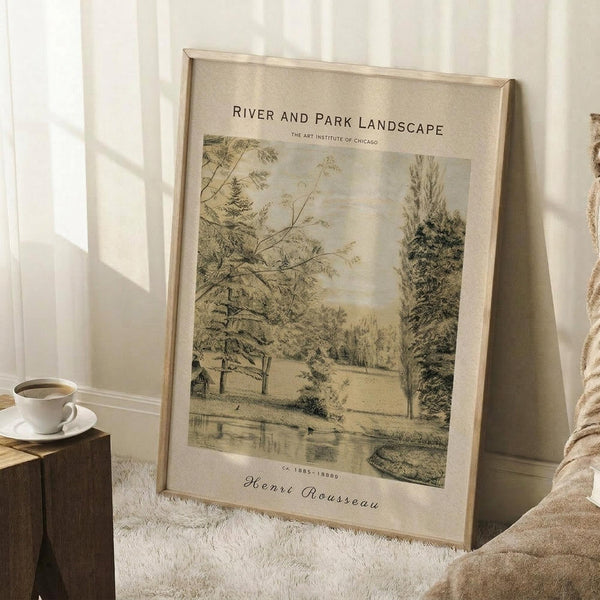 May include: A framed vintage-style landscape print leaning against a white wall corner, featuring trees, a river, and pastoral scenery in sepia tones. The print shows Art Institute of Chicago attribution and Henri Rousseau signature at bottom. A wooden side table holds a white coffee cup, with a cream knit throw blanket nearby on white carpet. Natural sunlight creates soft shadows on the wall.