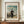 May include: Framed vintage art poster featuring a bearded man in dark clothing holding a painter's palette, standing in a whimsical landscape with bridge, buildings, and cloudy sky. The cream-colored print sits on a wooden surface beside a white ceramic vase and stacked books against light walls with white trim molding.