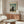 May include: vintage art poster of bearded man in black suit holding palette, standing before pastoral landscape with bridge and buildings under cloudy sky, displayed above mid-century walnut credenza in modern living room with cream chair, glass coffee table, and minimalist decor