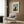 May include: Framed Henri Rousseau self-portrait print showing bearded man in dark suit holding palette against landscape with cloudy sky, displayed on wood paneled wall above cream upholstered armchair with geometric patterned pillow, brass floor lamp and dark side table with open book nearby