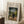 May include: Framed art print leaning against white paneled wall on textured cream rug. Print shows naive-style painting of man in dark suit and bowler hat standing next to small child in white dress in park setting with green trees. Museum-style layout with beige matting and thin wooden frame. Coffee cup on wooden block nearby.
