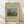 May include: Framed art print leaning against white wall on light wooden floor. Features Henri Rousseau's pastoral painting with brown cows grazing in green meadow, tall cypress trees, and large rounded tree. Cream-colored background with black text and 1910 date. Natural wood frame with visible grain. Soft shadows cast on textured white wall.