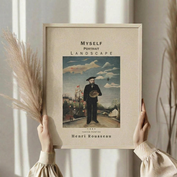 May include: Hands holding a framed art print featuring Henri Rousseau's self-portrait from 1890, showing a bearded man in black clothing holding a palette against a landscape with buildings and cloudy sky. The print has cream matting with text reading "MYSELF PORTRAIT LANDSCAPE" and "Henri Rousseau" in a light wooden frame. Dried pampas grass visible on the left side against a soft neutral background.