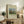 May include: Framed art print of Henri Rousseau's pastoral painting showing cattle grazing in green meadow with tall poplar trees and rounded tree. Cream matted poster in light wood frame sits on rustic wooden sideboard beside fern plant, stacked books, and ceramic vase in neutral-toned living room with beige armchair.