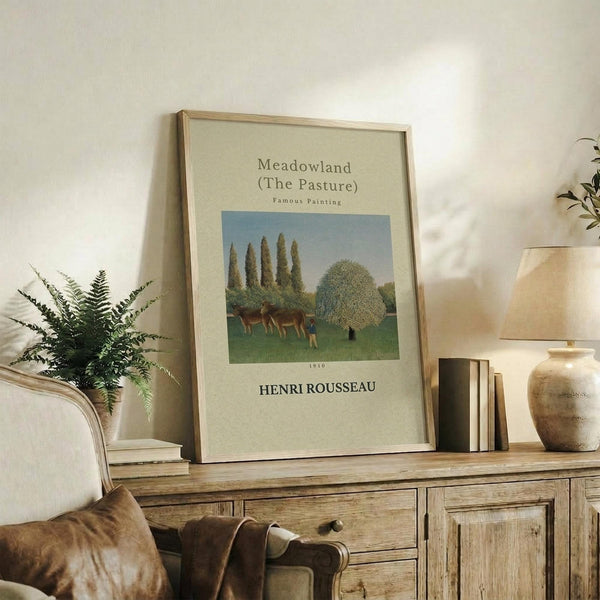 May include: Framed art print of Henri Rousseau's pastoral painting showing cattle grazing in green meadow with tall poplar trees and rounded tree. Cream matted poster in light wood frame sits on rustic wooden sideboard beside fern plant, stacked books, and ceramic vase in neutral-toned living room with beige armchair.