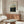 May include: Framed poster print featuring a lush forest scene with tall trees and dense foliage in greens and earth tones, displayed above a wooden sideboard with sliding doors in a modern living room setting. White sculptural chair and glass-top coffee table visible in foreground, with warm natural lighting creating shadows on cream walls.