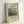 May include: Framed art print leaning against white wall showing Henri Rousseau's pastoral painting with brown cows grazing in green meadow, tall cypress trees, and rounded tree. Cream-colored background with black text identifying the artwork and artist. Natural wood frame visible from side angle.