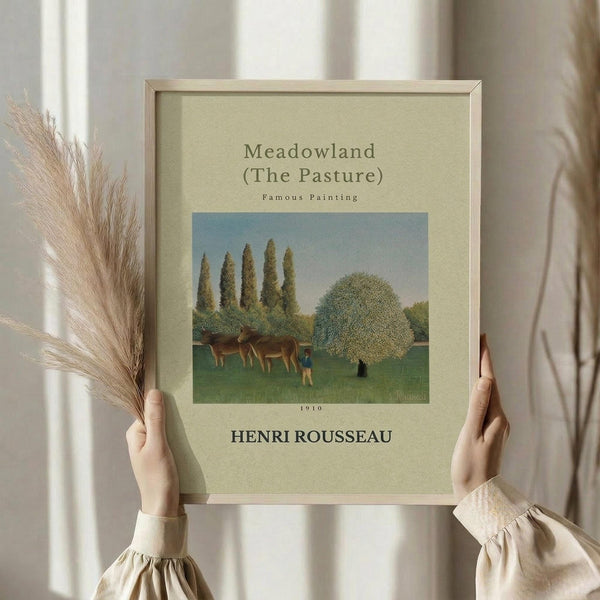 May include: Person in cream sweater holding framed art print featuring pastoral scene with cows grazing in green meadow, tall cypress trees, and large leafy tree under blue sky. Print has cream matting in light wood frame. Dried wheat stems visible on left side against soft curtained background.
