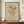 May include: Framed botanical illustration print displayed on wooden credenza in elegant room with cream walls and decorative molding. The vintage-style artwork features hand-drawn flowers and plants in sepia tones on aged paper background with cursive text below. White ceramic vase and books sit nearby. Ornate wall sconce visible on right side.