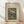 May include: Framed vintage botanical poster showing colorful flowers in a glass vase against green background, displayed on light wooden floor against white wall with natural lighting and shadows. The artwork features pink peonies, white daisies, and foliage in muted tones with cream matting and natural wood frame.