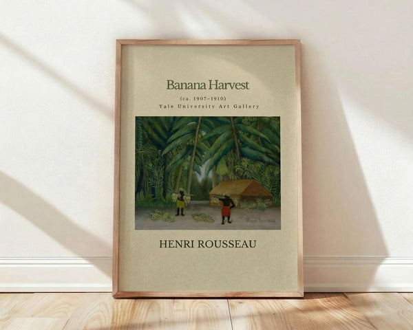 May include: Framed art print leaning against white wall on light wood floor. Cream-colored poster features tropical jungle scene with banana trees, small figures harvesting beneath large green foliage. Natural wood frame with thin profile. Soft natural lighting creates diagonal shadows across white textured wall. Print shows museum-style layout with beige background and black text above and below central image.