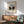 May include: framed art print of a tree-lined canal waterway in muted greens and browns, displayed above a wooden credenza with white ceramic lamp, next to a mint green upholstered mid-century armchair with wooden frame, in a bright room with natural sunlight shadows on white walls