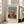 May include: Framed art print leaning against beige sofa in bright living room with white walls and windows. Print features naive-style painting of blonde child in red polka dot dress holding small doll, seated against blue and green background. Text reads "Child with Doll" and "Famous Painting Henri Rousseau (1906)" on cream colored border around artwork.