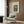 May include: Vintage botanical poster print titled "Flowers in a Vase" by Henri Rousseau displayed in a modern reading nook. The artwork features pink carnations and other flowers in a glass vase against a green background, framed in light wood and hung on wood-paneled walls. Scene shows a cream upholstered armchair with geometric pillow, brass floor lamp, and dark round side table with open book.