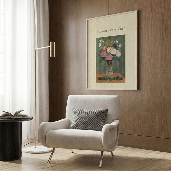 May include: Vintage botanical poster print titled "Flowers in a Vase" by Henri Rousseau displayed in a modern reading nook. The artwork features pink carnations and other flowers in a glass vase against a green background, framed in light wood and hung on wood-paneled walls. Scene shows a cream upholstered armchair with geometric pillow, brass floor lamp, and dark round side table with open book.