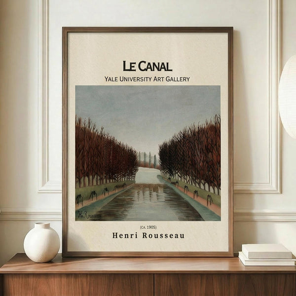 May include: Framed art print of Henri Rousseau's "Le Canal" painting showing a tree-lined waterway with bare winter branches creating a natural archway over a reflective canal. The poster features muted earth tones and is displayed in a thin brown frame on a wooden sideboard alongside a white ceramic vase and stacked books in a minimalist room with cream walls and white molding.