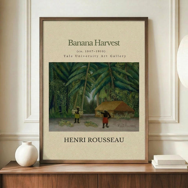 May include: Framed art print of tropical jungle scene with banana trees and two small figures beneath thatched shelter, displayed on wooden credenza next to white ceramic vase and stacked books against cream colored wall with white trim molding.