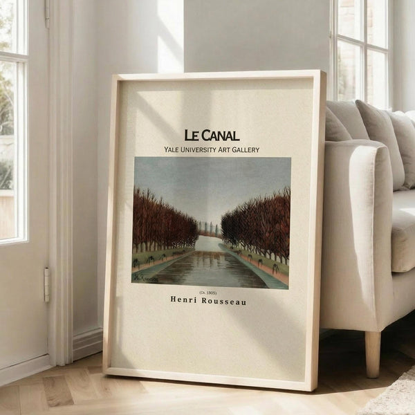 May include: Framed art print featuring a serene canal scene with autumn trees lining both sides, painted in muted greens and browns with a pale sky. The artwork is displayed in a light wooden frame, leaning against a white wall next to a beige upholstered chair in a bright, sunlit room with large windows and hardwood floors.