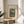 May include: Framed vintage botanical poster featuring colorful flowers in a vase against green background, displayed on round wooden side table next to beige upholstered chair. Soft natural lighting creates shadows across cream colored wall. Plant leaves visible in corner of minimalist, neutral-toned room setting.