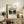 May include: Framed art print displaying a canal scene with tree-lined waterway in muted greens and browns, positioned on rustic wooden sideboard alongside fern plant, stacked books, and ceramic vase in neutral-toned living room setting with beige armchair and white walls
