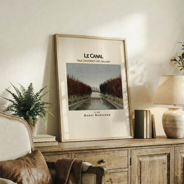 May include: Framed art print displaying a canal scene with tree-lined waterway in muted greens and browns, positioned on rustic wooden sideboard alongside fern plant, stacked books, and ceramic vase in neutral-toned living room setting with beige armchair and white walls