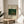 May include: A framed art print displayed on a round wooden side table next to a beige upholstered chair. The poster features a tropical jungle scene with lush green palm trees and foliage, small figures harvesting bananas, rendered in a naive art style. The print has cream-colored matting with text above and below the artwork, housed in a thin gold frame. Soft natural lighting creates gentle shadows, with a potted plant visible in the left corner of the minimalist, neutral-toned room setting.