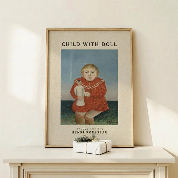May include: Framed art print displaying Henri Rousseau's naive-style painting of a blonde child in a red polka-dotted dress holding a small doll, set against blue and green background. Light wood frame with cream matting, displayed on pale wooden shelf alongside small wrapped gift with twine and evergreen sprig. Soft natural lighting on neutral wall.