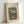 May include: Framed vintage botanical art print featuring colorful flowers in a ribbed glass vase against green background. Shows pink peonies, white daisies, tulips and foliage in Henri Rousseau's distinctive style. Natural wood frame with white mat, leaning against white wall corner on light wooden surface.