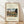 May include: Framed poster print of Henri Rousseau's 1910 painting showing the Eiffel Tower and Seine River at sunset, featuring naive art style with vibrant autumn colors, bridge, and lush greenery. Light wooden frame with cream matting, leaning against white wall on light wood flooring with soft natural lighting creating diagonal shadows.