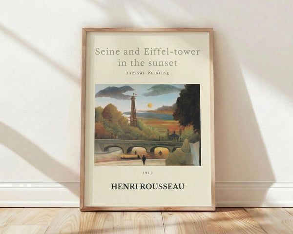 May include: Framed poster print of Henri Rousseau's 1910 painting showing the Eiffel Tower and Seine River at sunset, featuring naive art style with vibrant autumn colors, bridge, and lush greenery. Light wooden frame with cream matting, leaning against white wall on light wood flooring with soft natural lighting creating diagonal shadows.