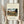 May include: Hands holding framed art print featuring Eiffel Tower and bridge scene in autumn colors with yellow sun, displayed against white curtained window with dried pampas grass. Cream matted print in light wood frame shows naive style painting with green foliage, stone bridge arches, and small figure by water.